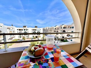 Een balkon met een tafel, overdekt met fruit en een glas sap, met uitzicht op een woongebied met witte gebouwen en een heldere lucht.