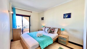 A bedroom with a bed, a nightstand, a green rug, a window with curtains, and a painting on the wall. - ALBUFEIRA SALGADOS BEACH 6 by HOMING (Albufeira)
