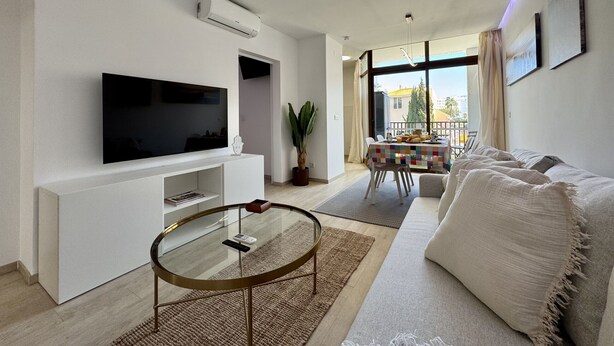 Una sala moderna con televisor de pantalla plana, una mesa de centro de vidrio, un sofá y un balcón con mesa y sillas.