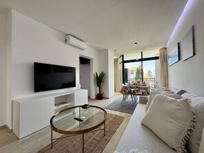 Ein modernes Wohnzimmer mit Flachbildfernseher, Glastisch, Sofa und einem Balkon mit Tisch und Stühlen.