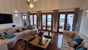 En moderne stue med sofa, sofabord og fjernsyn. Store vinduer med gardiner, og der er synlig en balkon gennem vinduerne.