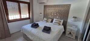 Ein Schlafzimmer mit einem Bett, zwei Nachttischen, einem dekorativen Kopfteil und einem Fenster mit Jalousien.