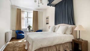 A bedroom with a large bed, a side table, a chair, a window with curtains, and a radiator.