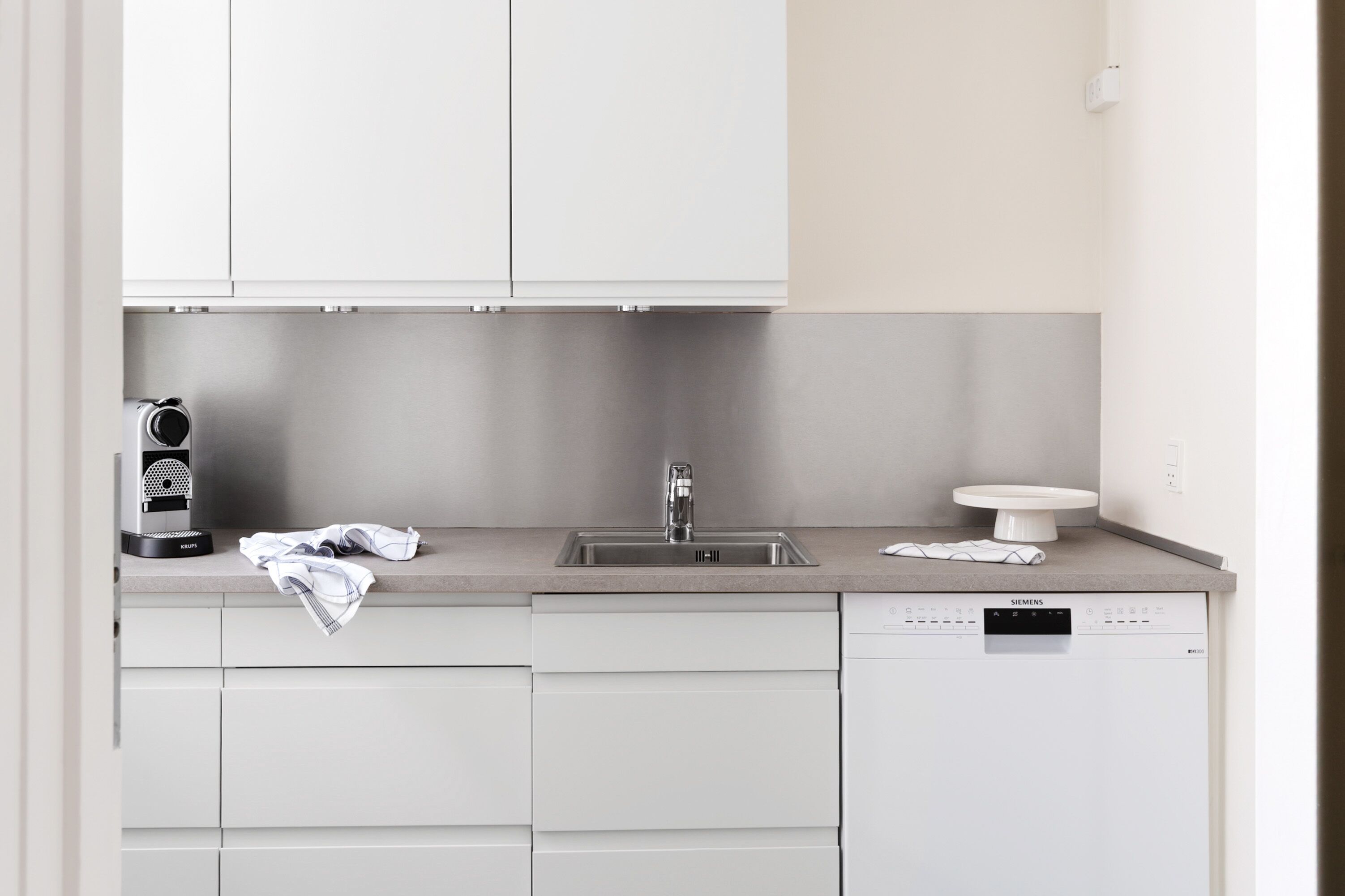 Uma cozinha moderna com armários brancos, uma pia de aço inoxidável, uma lava-louças Siemens e uma cafeteira.