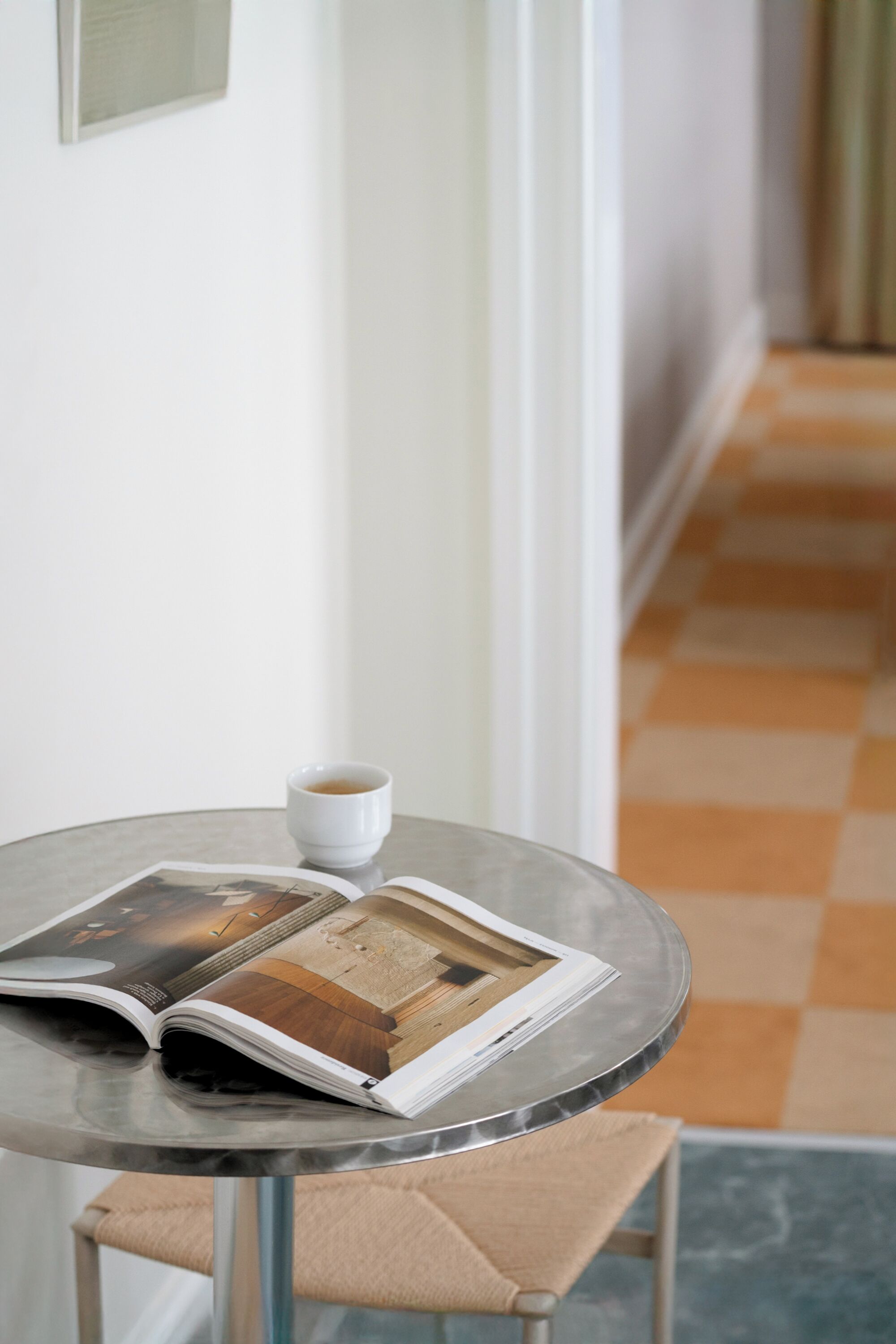 Una mesa redonda con una revista, una taza de café, una silla y un suelo a cuadros.