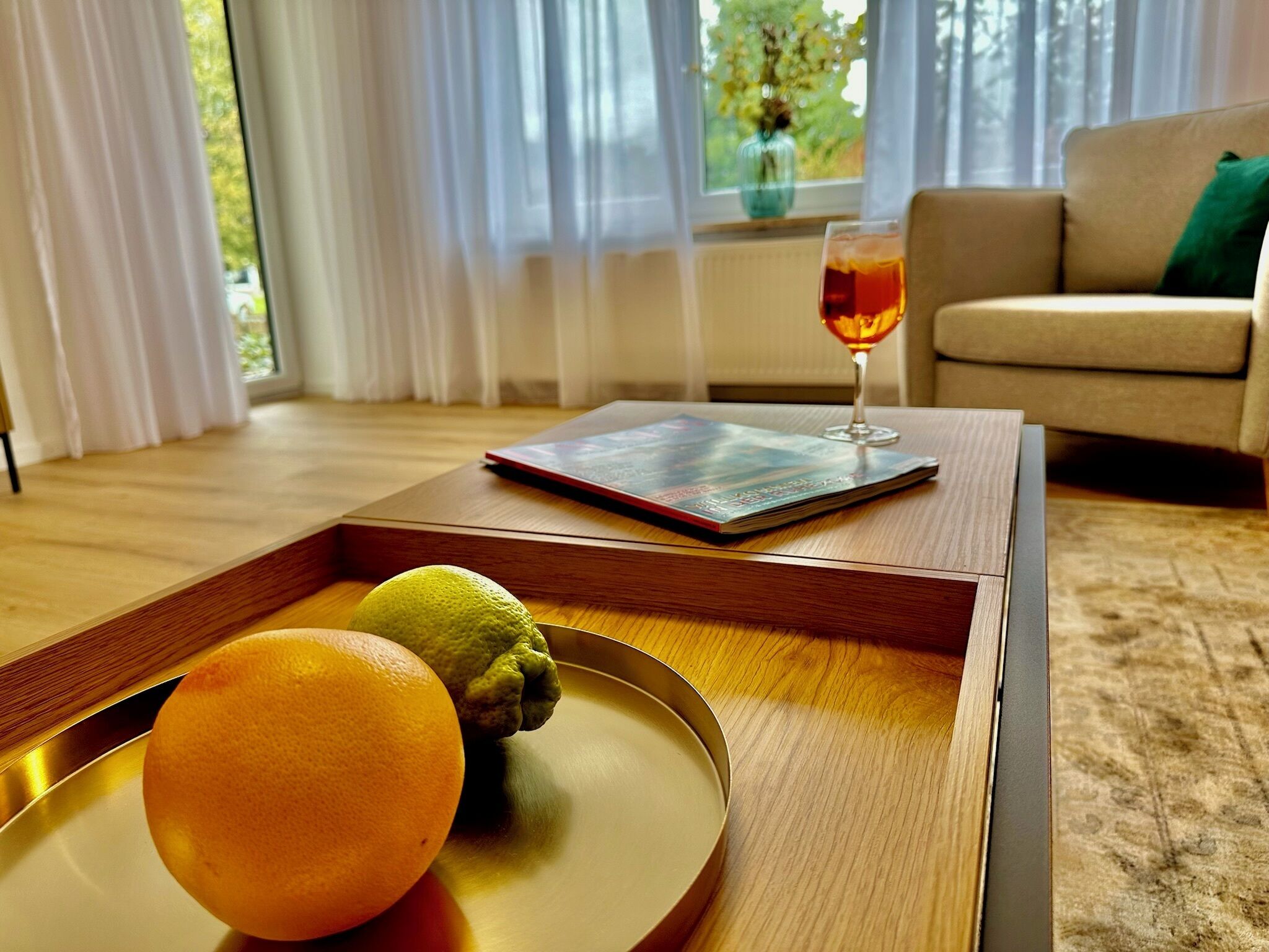 Uma bandeja de madeira com um limão e uma laranja sobre uma mesa, uma revista e um copo de bebida vermelha em uma sala de estar com sofá e janela com cortinas.