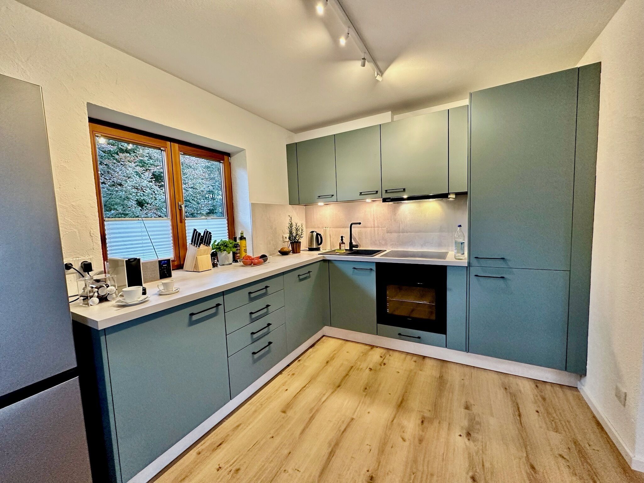 A modern kitchen with a wooden floor, light blue cabinets, and a window with a view of trees.