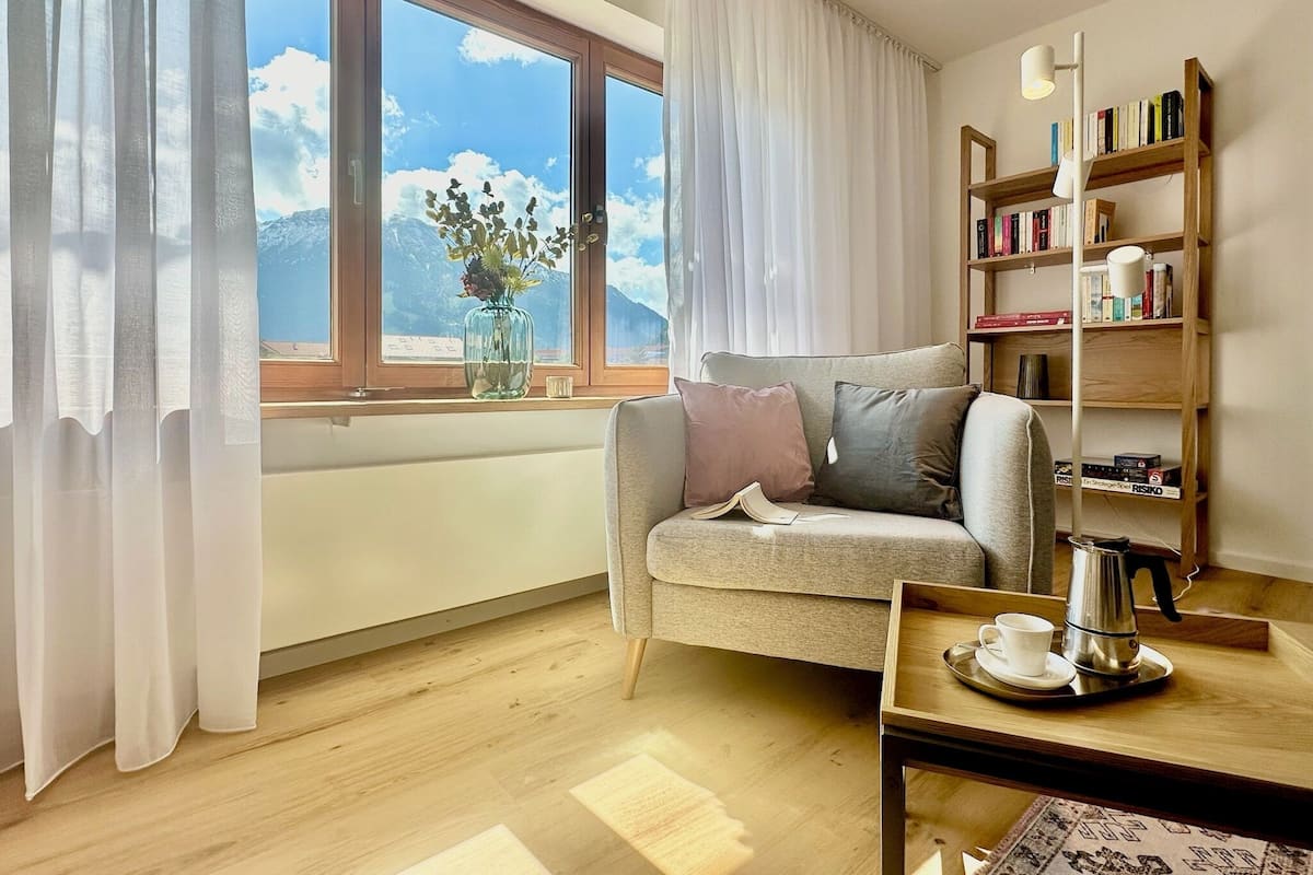 A cozy living room with a sofa, a coffee table, a bookshelf, and a window with a view of mountains.