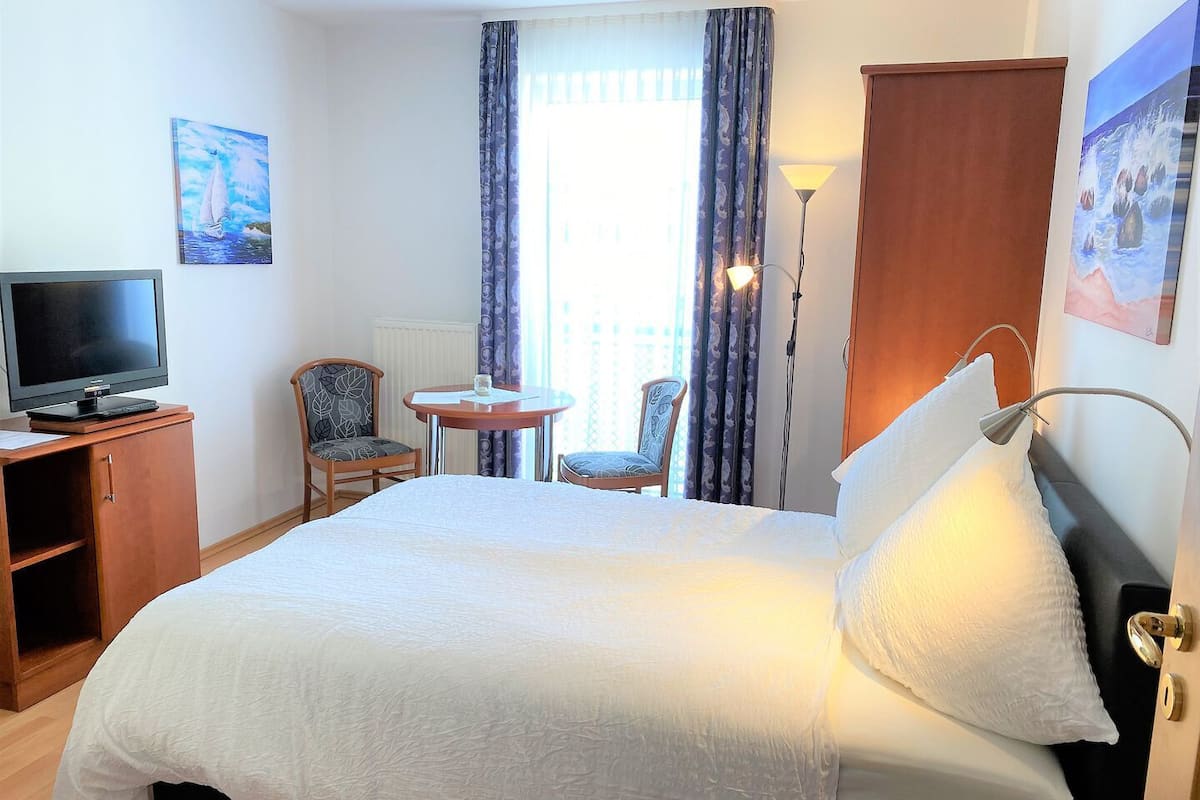 A hotel room with a bed, a desk with a TV, two chairs, a small table, and a window with curtains.