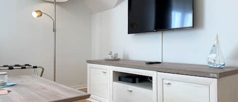A modern living room with a flat-screen TV mounted on the wall, a wooden dining table with chairs, a small cabinet with drawers, and a floor lamp.