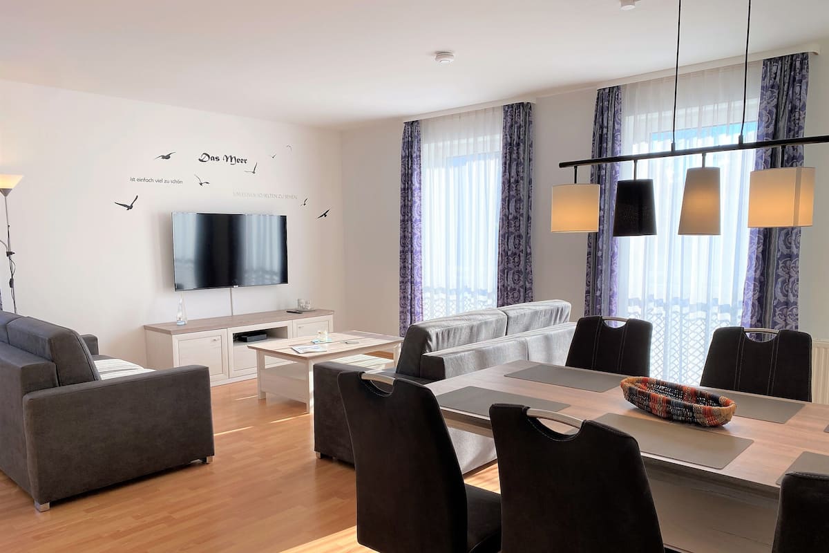 A modern living room with a sofa, a dining table with chairs, a flat-screen TV, and a large window with curtains.