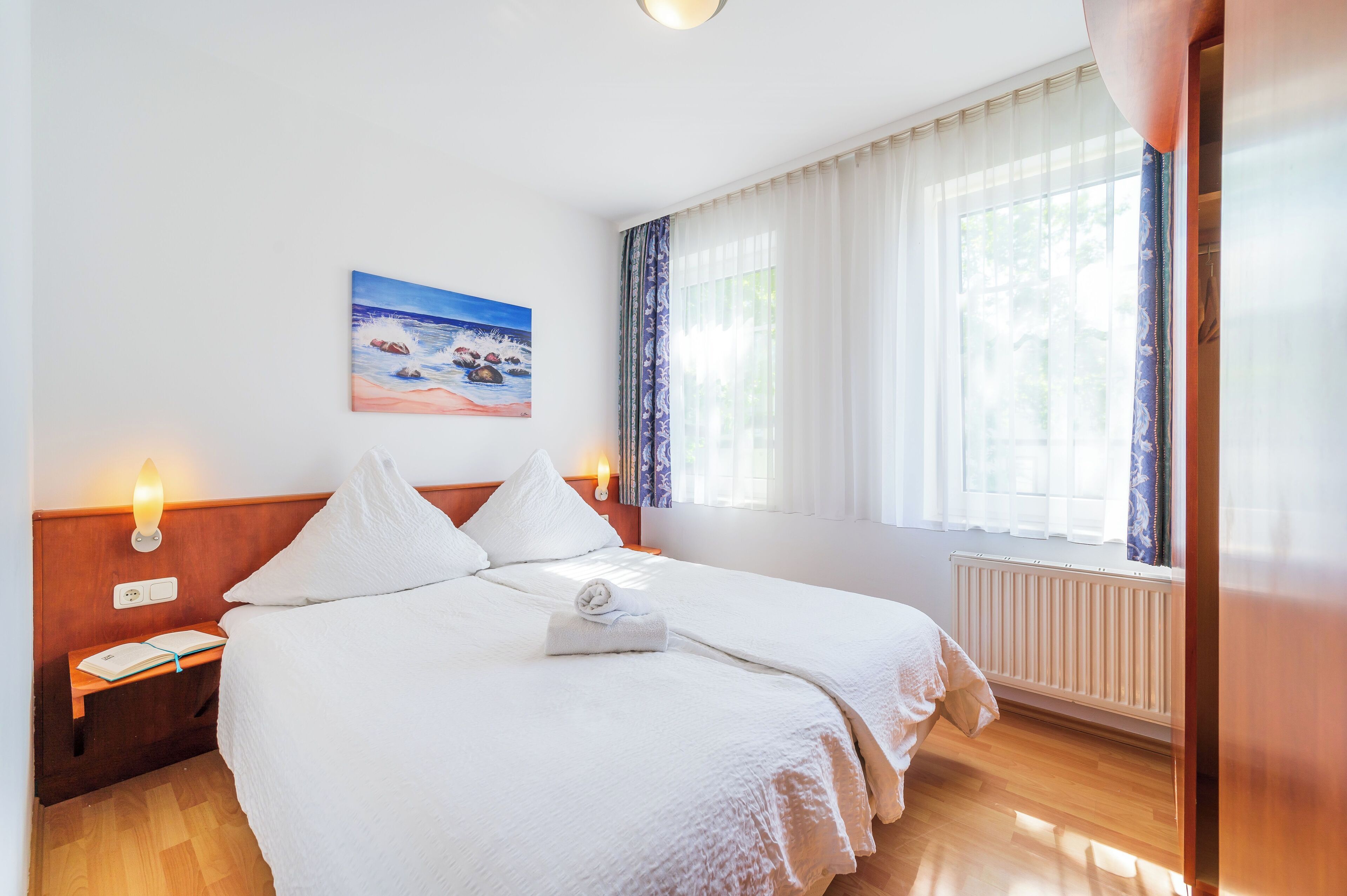 Habitación de hotel con dos camas, un cuadro de una escena de playa, una mesita de noche con un libro y una lámpara, y una ventana con cortinas traslúcidas.