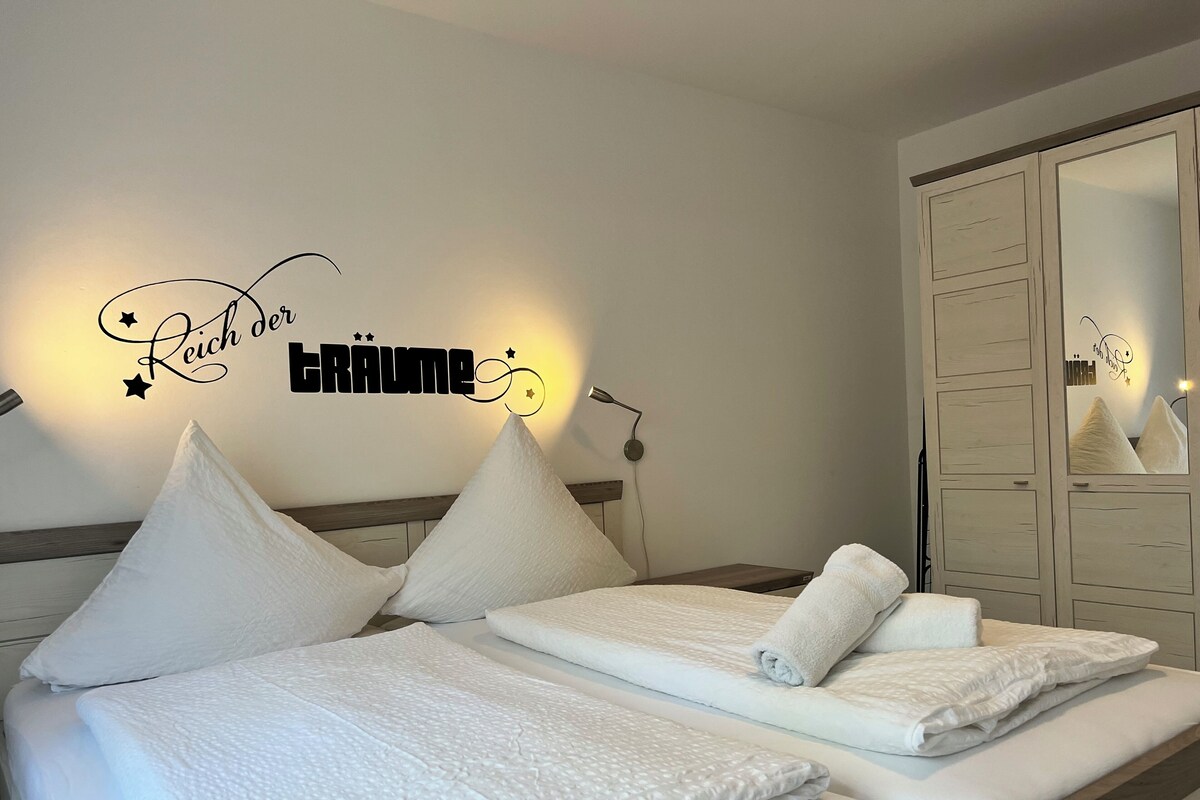 A neatly made bed with white linens and pillows, a bedside lamp, and a wall decoration with text and stars.
