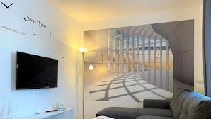 A modern living room with a large flat-screen TV, a sofa, a small coffee table, and a wall mural depicting a sunlit corridor.