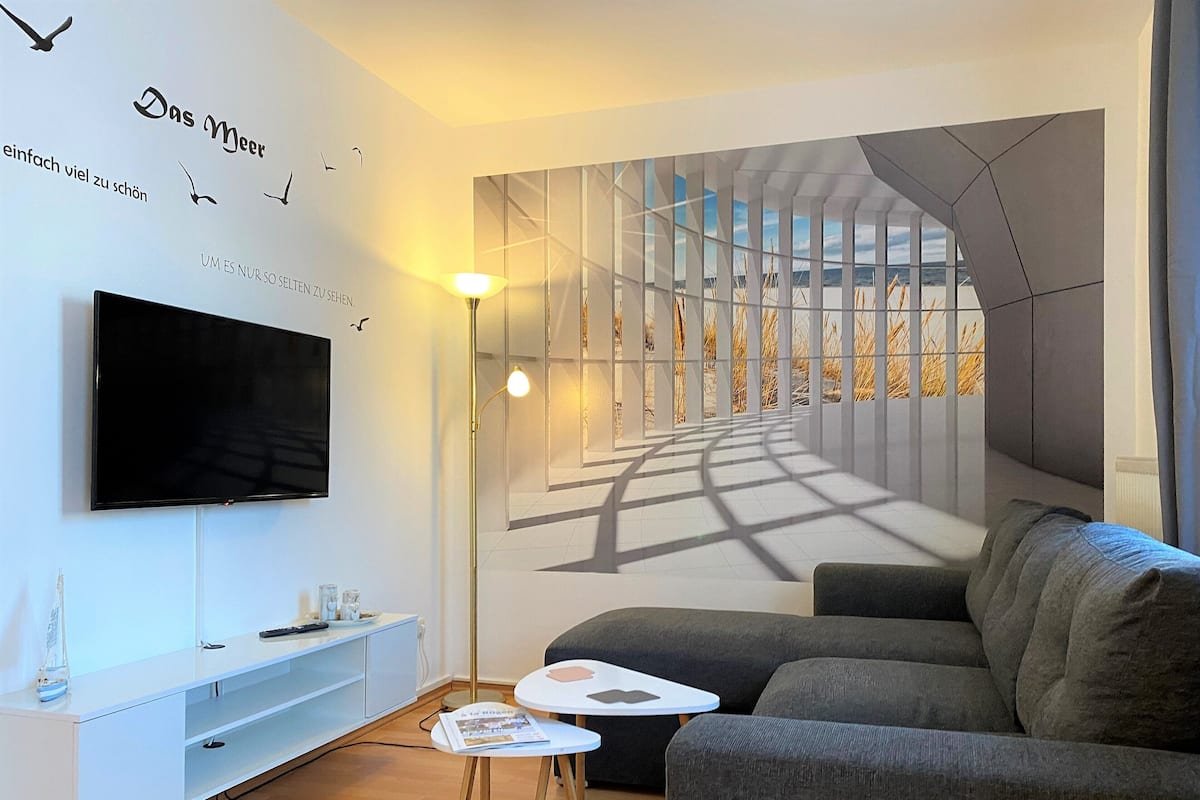 A modern living room with a large flat-screen TV, a sofa, a small coffee table, and a wall mural depicting a sunlit corridor.