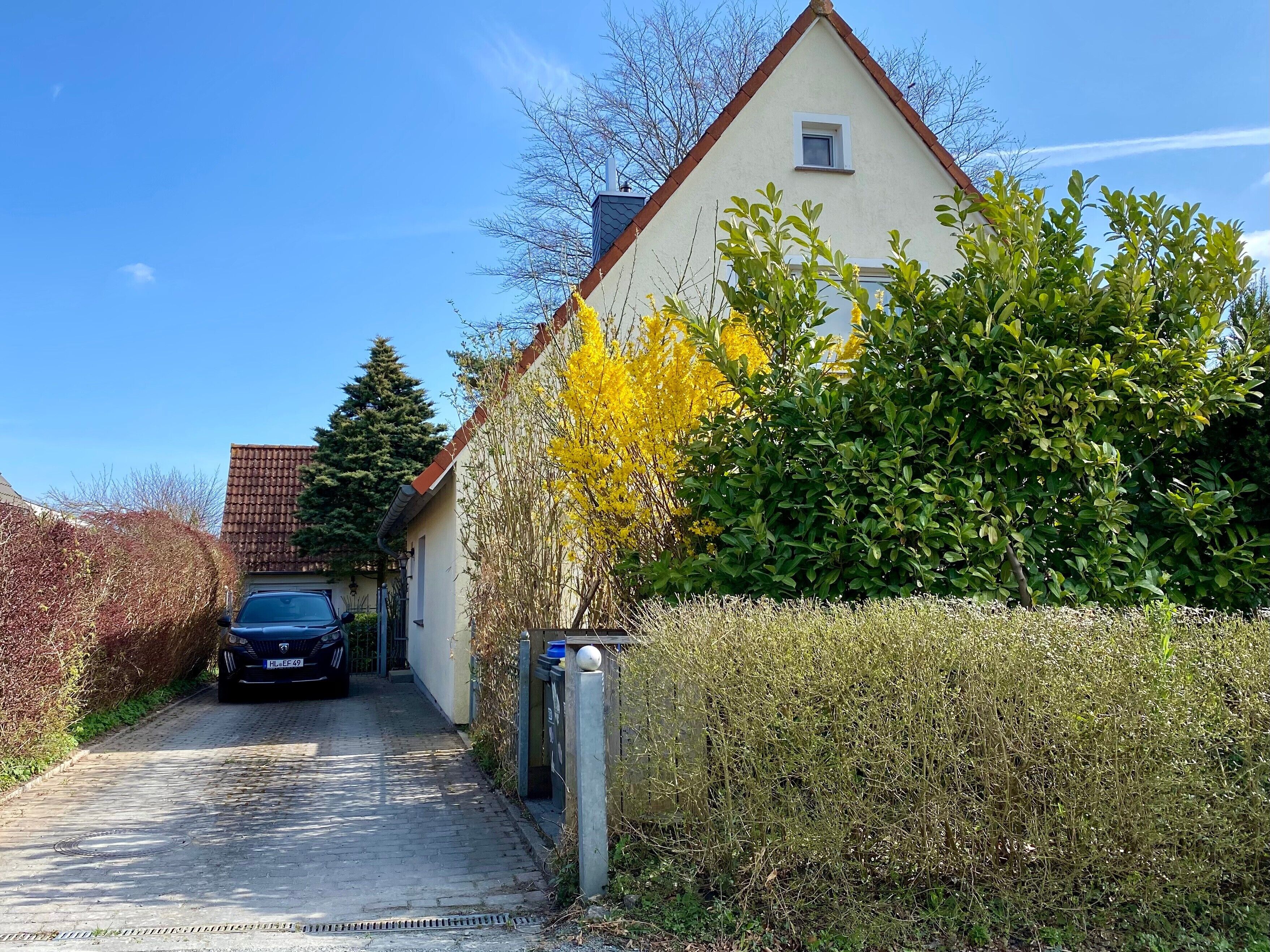 A residential house with a paved driveway, a parked car, and a hedge in front.