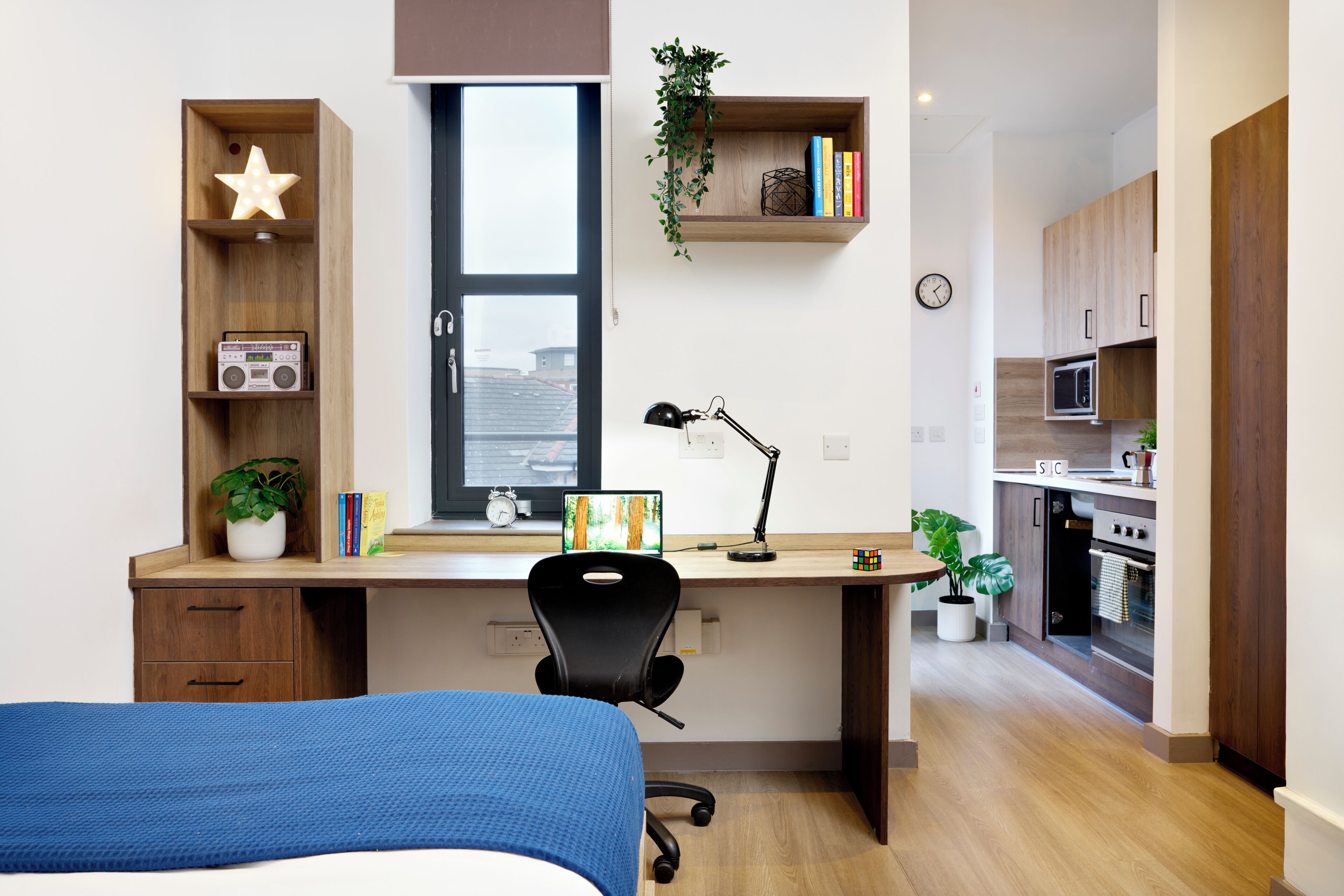 Ein modernes Schlafzimmer mit einem Bett, einem Schreibtisch mit Stuhl, einem Fenster und einer Küchenzeile.