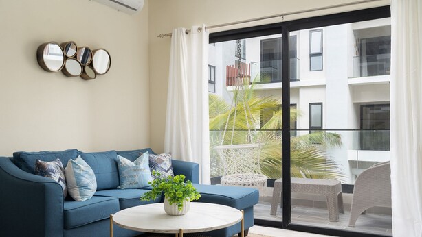 A modern living room with a blue sofa, a round coffee table, and a large window with a view of a building and palm trees. - Cozy 1BR in Panorama Lake • Pool & Beach (PUNTA CANA)