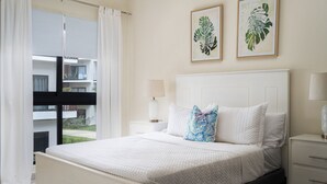 A bedroom with a bed, white curtains, a bedside table with a lamp, and two framed plant prints on the wall. - Cozy 1BR in Panorama Lake • Pool & Beach (PUNTA CANA)