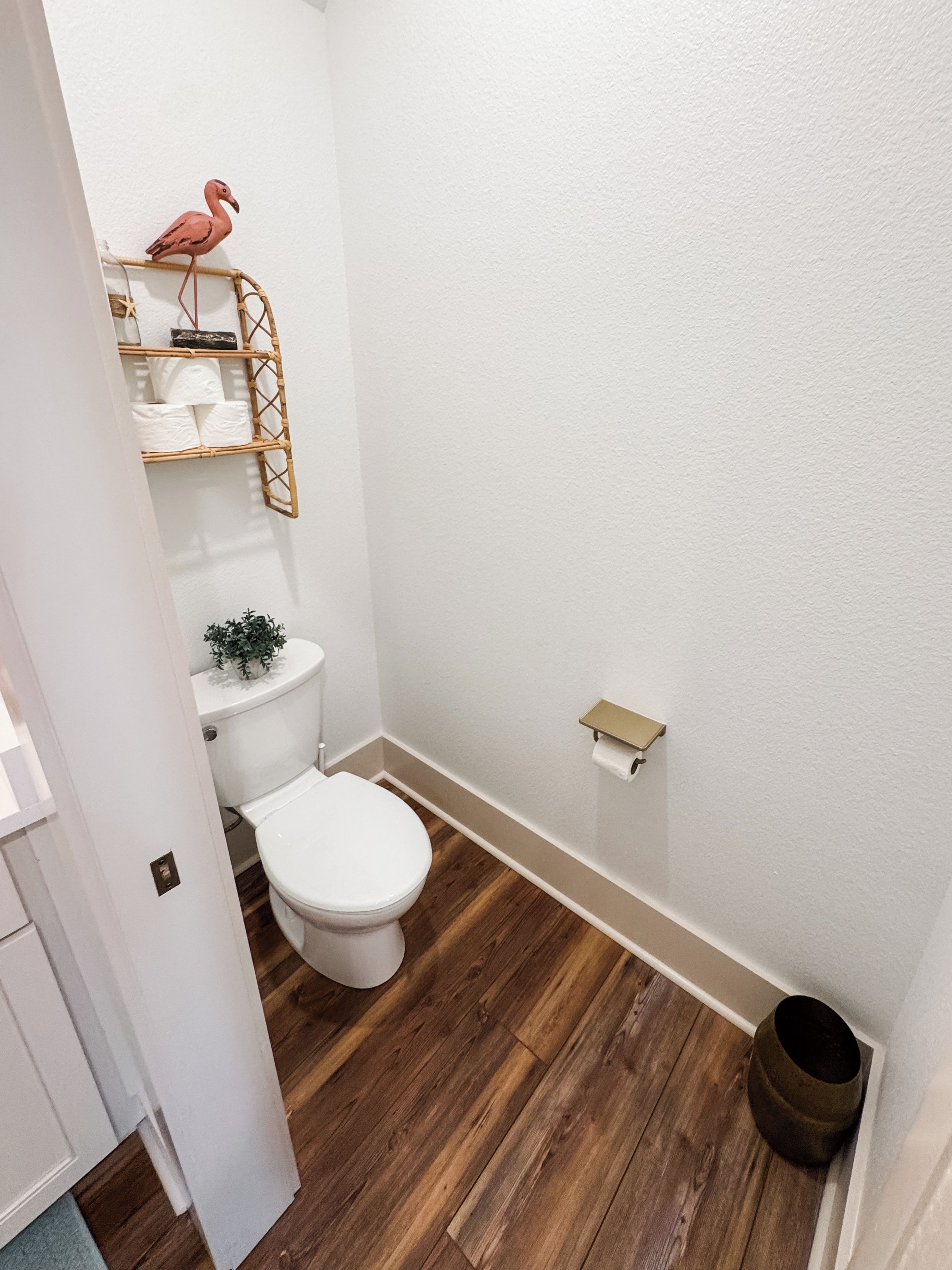 A small bathroom with a white toilet, wooden flooring, a white wall, a small shelf with a pink flamingo decoration, and a trash can.