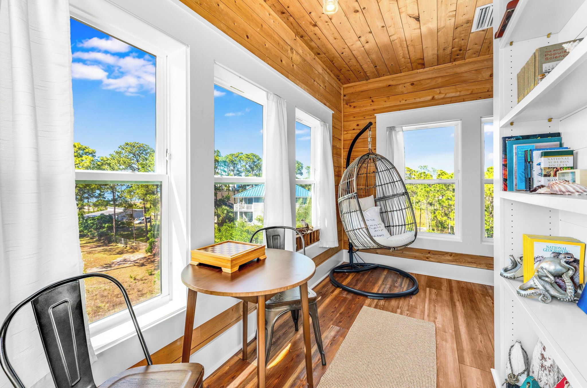 A room with large windows, a wooden ceiling, a round table with a chessboard, a hanging chair, and bookshelves.
