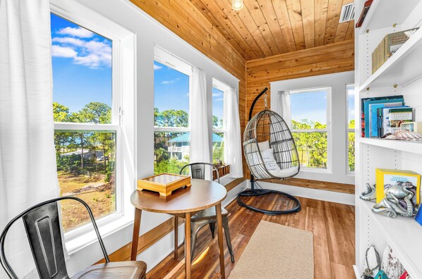 A room with large windows, a wooden ceiling, a round table with a chessboard, a hanging chair, and bookshelves.