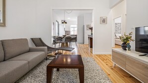 Una sala de estar moderna con un sofá, una mesa de centro y un televisor sobre un mueble. En el fondo, se observa un área de comedor con una mesa y sillas.