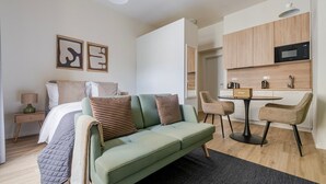 Une chambre moderne et bien éclairée, avec un canapé vert, un lit agrémenté d’oreillers à rayures et une petite kitchenette équipée d’un four à micro-ondes et d’une machine à café.