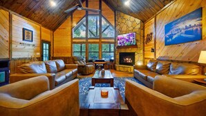Ein gemütliches Blockhäuschen-Interieur mit Ledersofas, Kamin, Fernseher und einem großen Fenster.