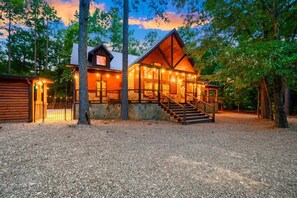 A rustic cabin with a wooden exterior, a stone foundation, and a staircase leading to a porch with string lights. - After the Sunset! 1-Level 5BR! Pool! - Arcade! (Broken Bow)