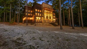 A two-story log cabin with a stone foundation, surrounded by tall pine trees.