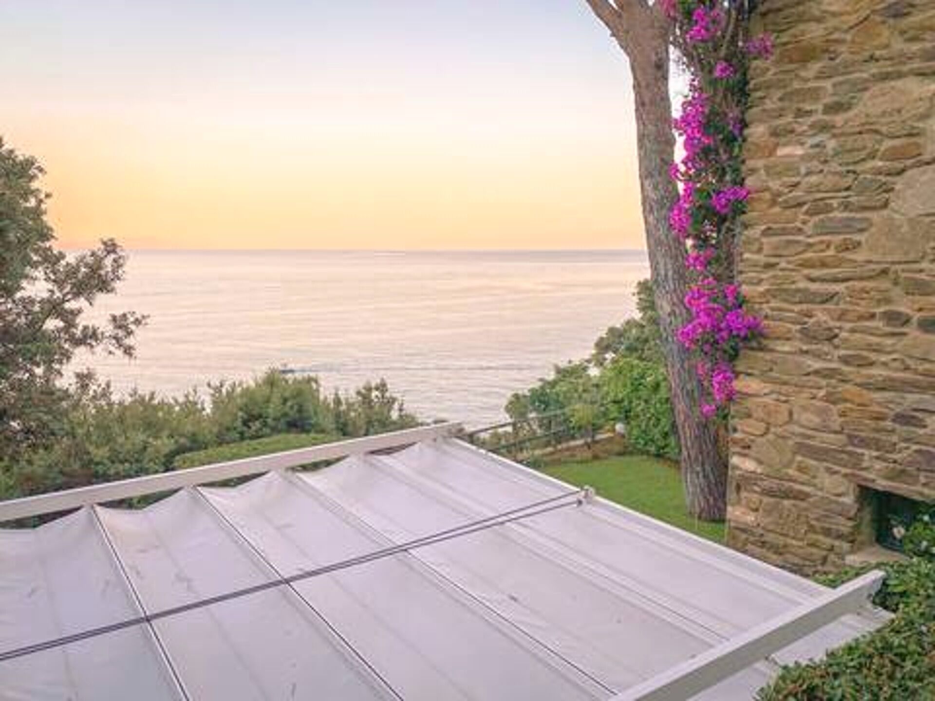 Une vue d’une zone côtière avec un bâtiment en pierre, un arbre aux fleurs roses et une structure blanche au premier plan.