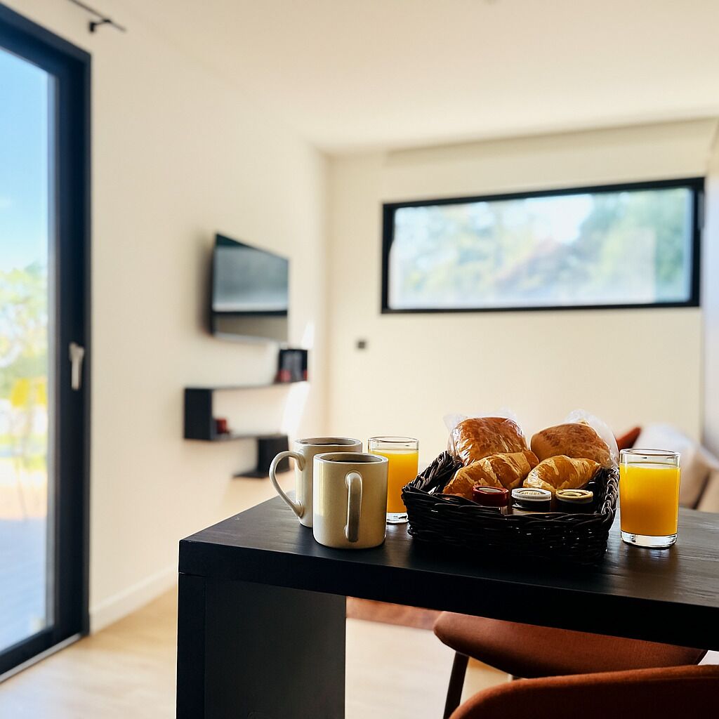A breakfast setup with croissants, coffee, and orange juice on a table in a room with a large window, a TV, and a bookshelf.