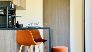 A modern kitchen with a built-in oven and microwave, two orange chairs, a small round ottoman, and a dining table with a plate and glasses. - Domaine du Mafay - Provençal Lodge (Bourg-des-Comptes)