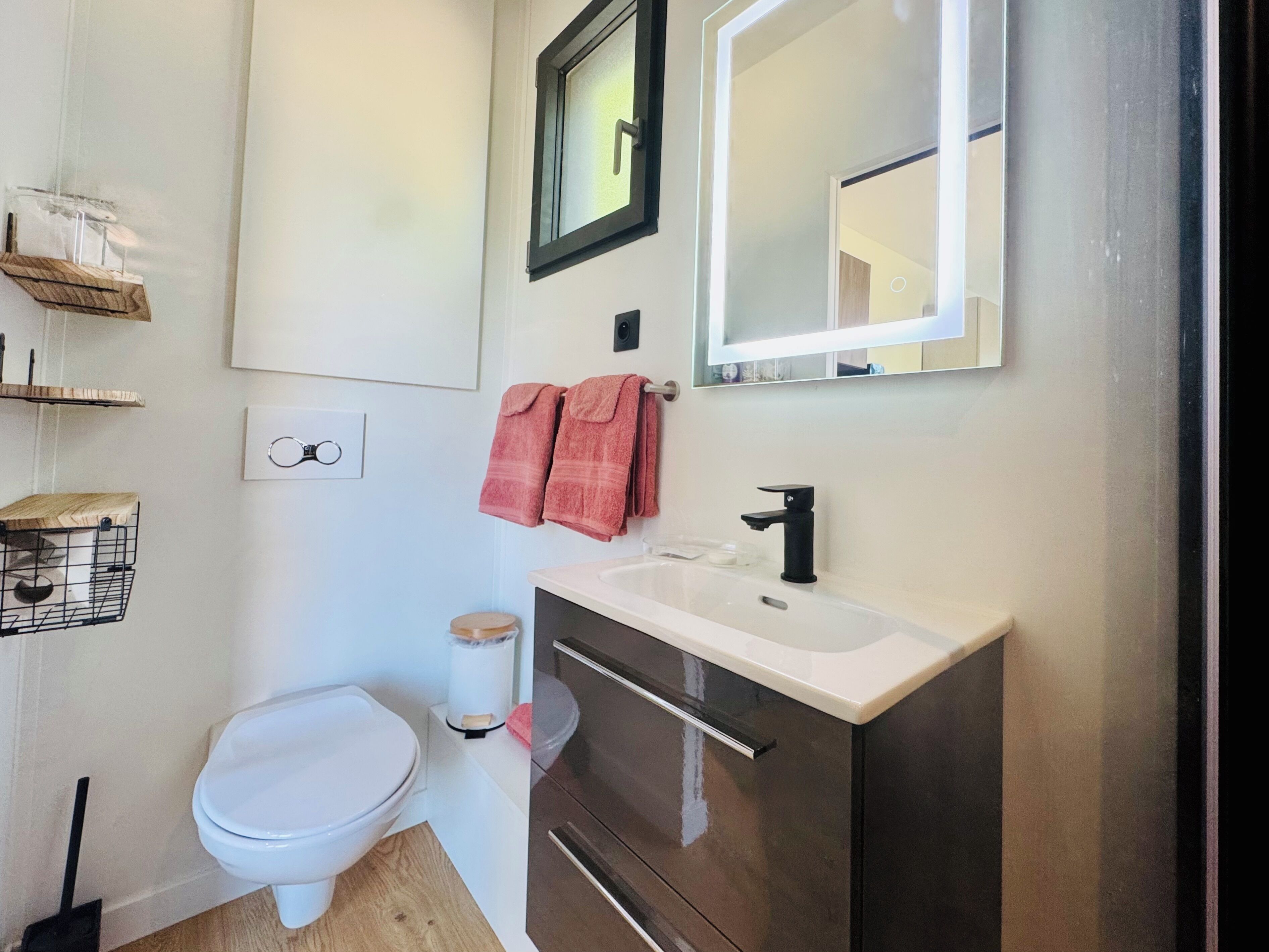 A modern bathroom with a white toilet, a vanity with a black faucet, and a mirror above the sink. There are two towels hanging on the wall and a small shelf with a basket.