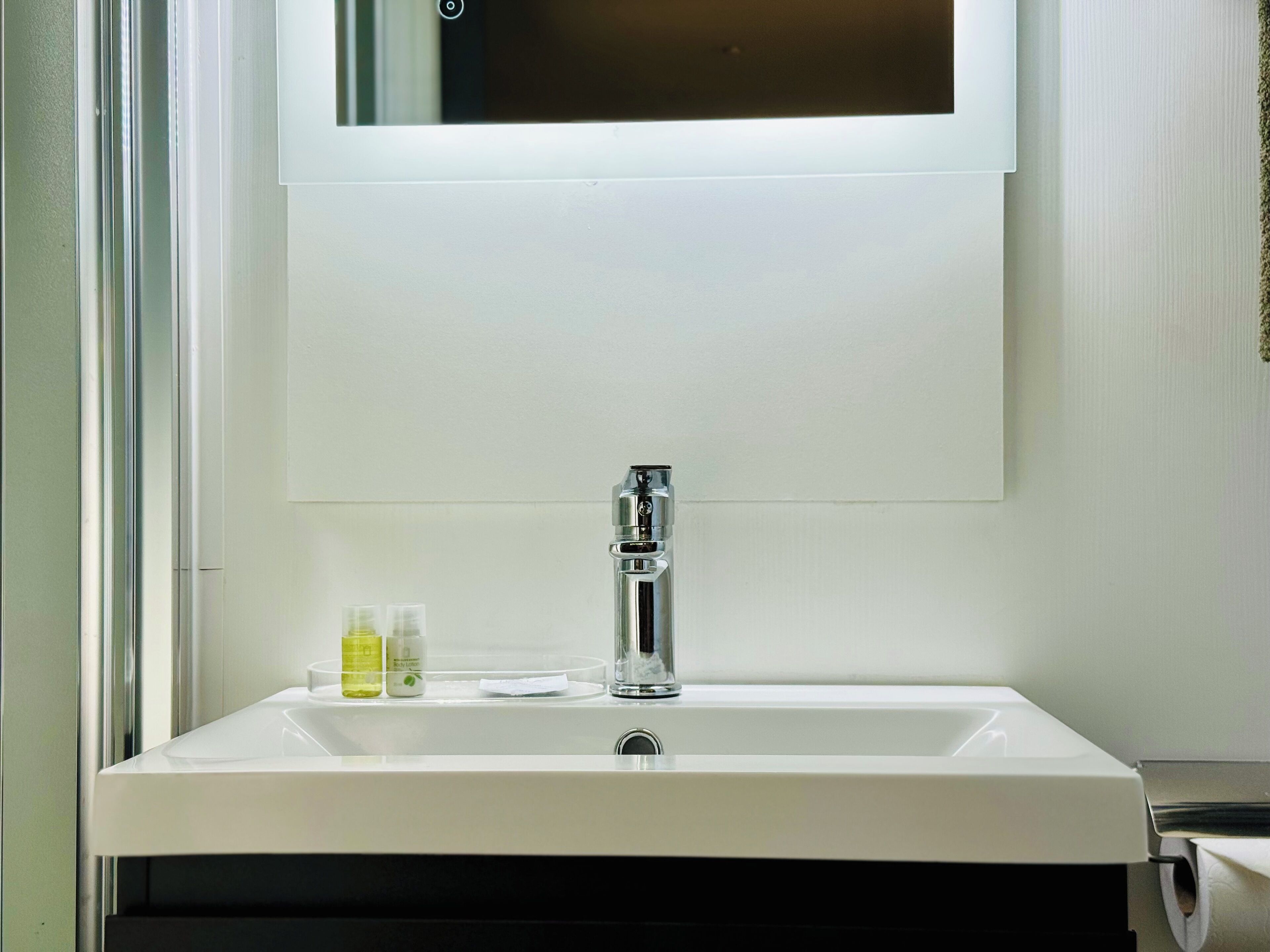 Un lavabo bianco con rubinetteria cromata, uno specchio sopra e due piccole bottigliette sul piano.