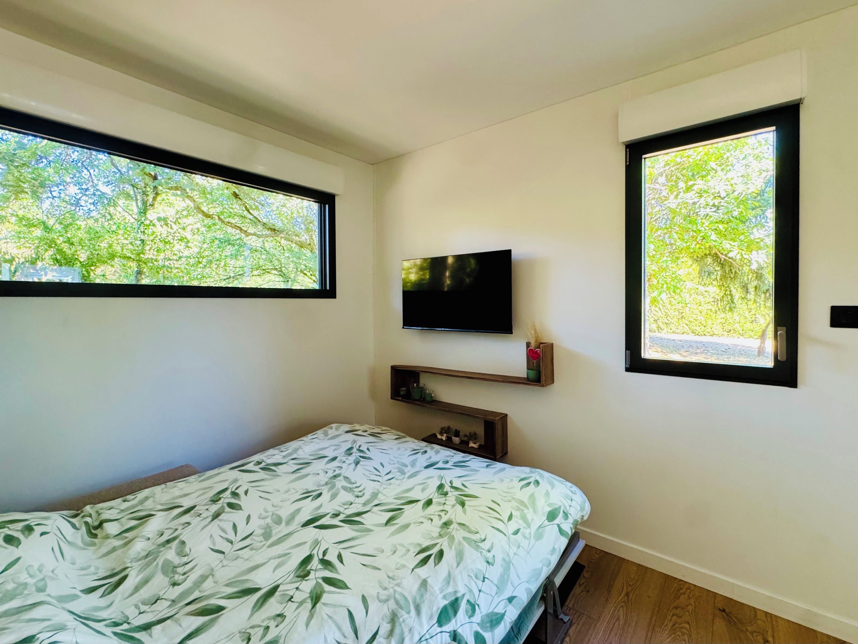 Quarto com cama, televisão presa na parede e duas janelas com vista para área verde.