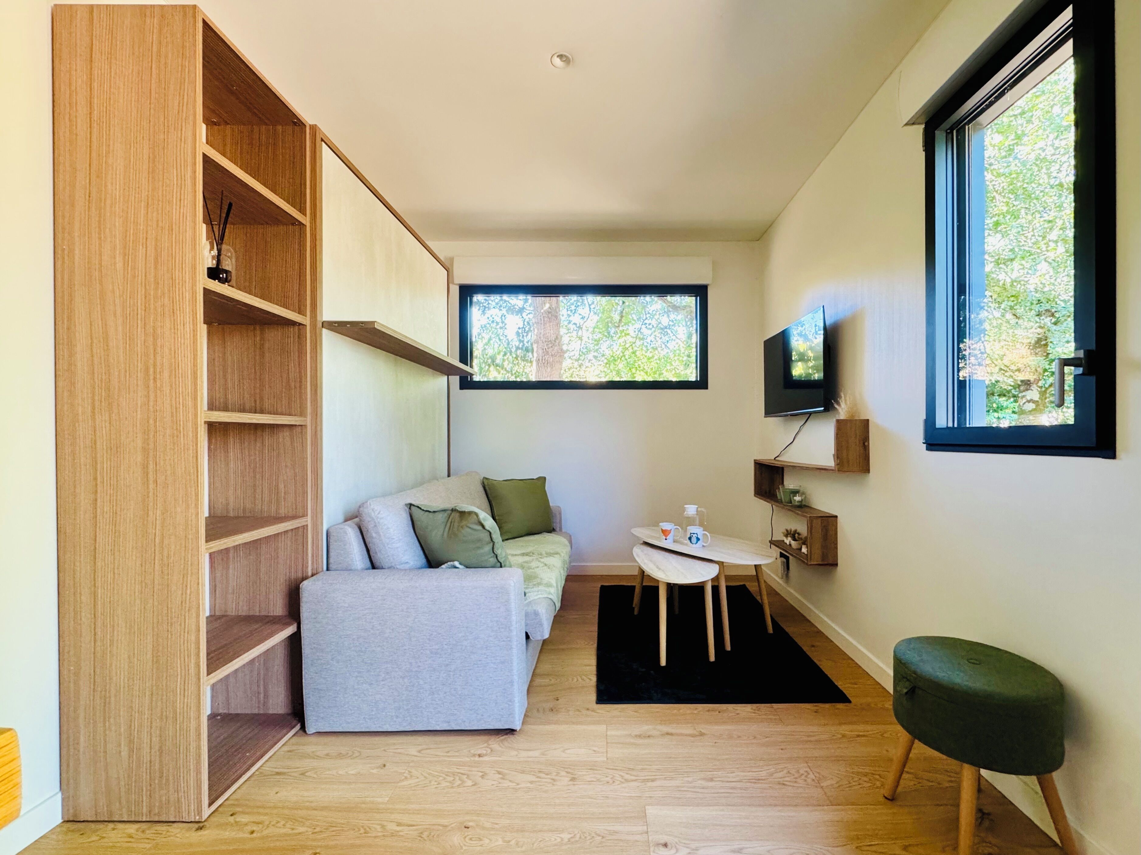Een knus leefruimte met een ingebouwde boekenkast, een grijze bank met kussens, een tafeltje met een stoel, een aan de muur bevestigde televisie en een raam met uitzicht op bomen.
