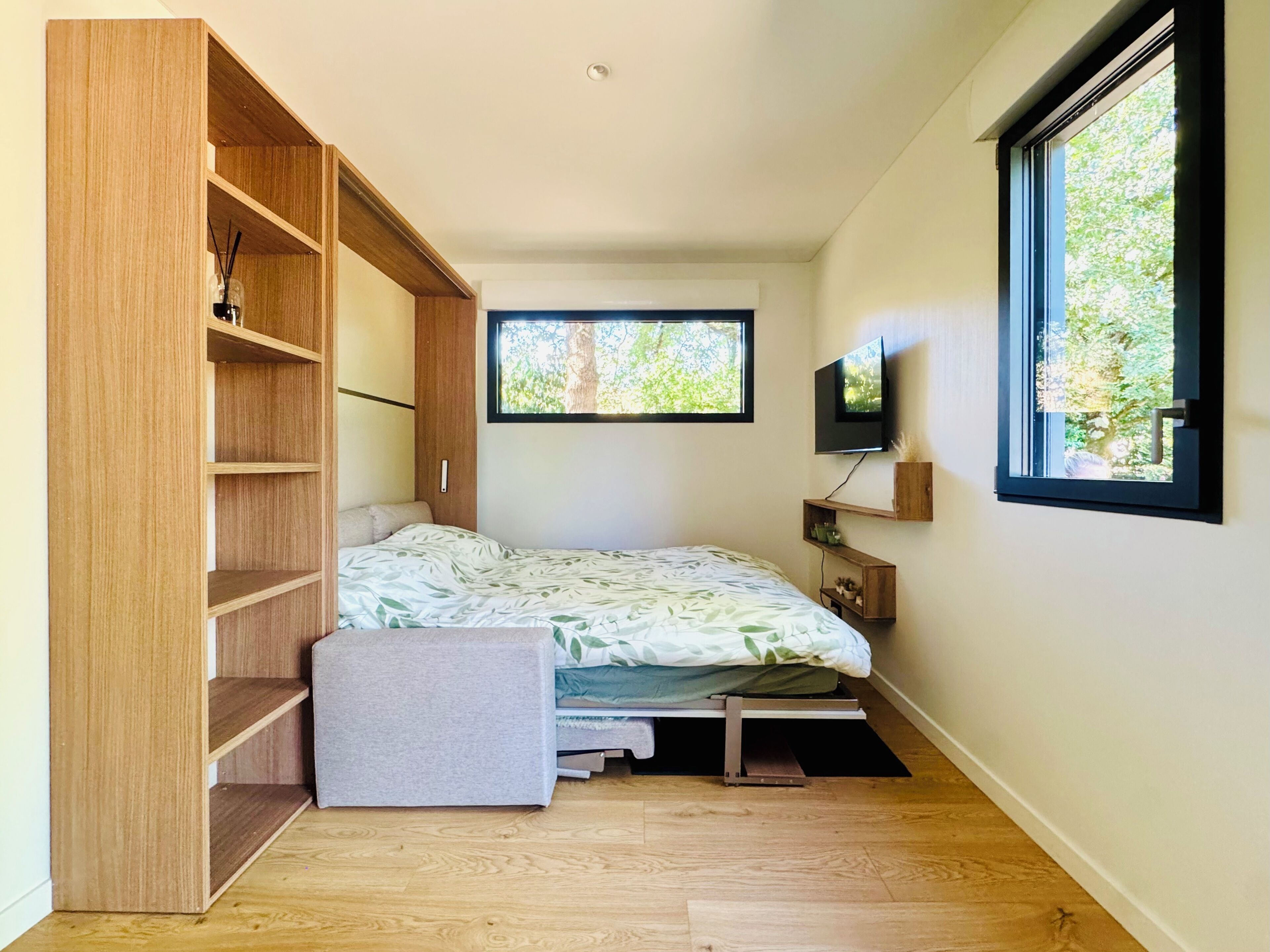 Une chambre à coucher de taille modeste, comprenant un lit, des étagères en bois intégrées, un téléviseur et une fenêtre donnant sur un arbre.