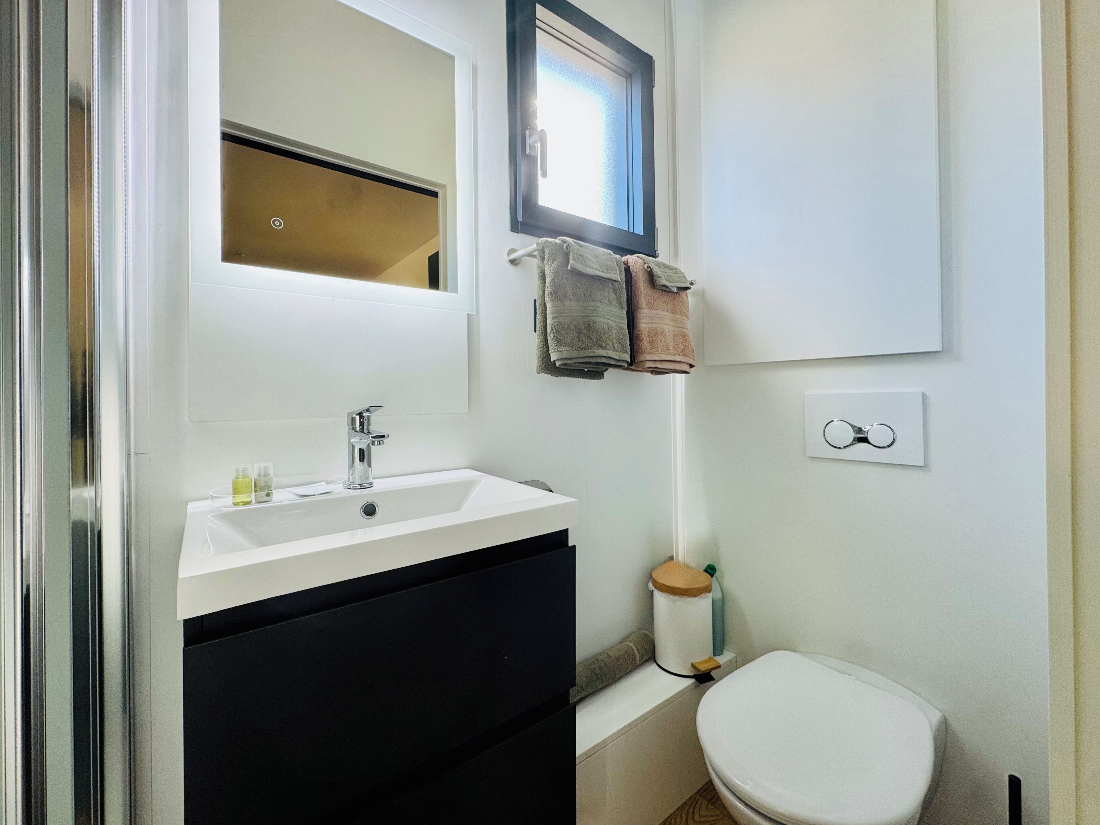 Une salle de bain avec un lavabo blanc, un miroir au-dessus, un porte-serviettes avec des serviettes, des toilettes et une petite fenêtre.