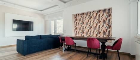 A modern living room with a blue sofa, a dining table with red chairs, and a large abstract wall art piece.