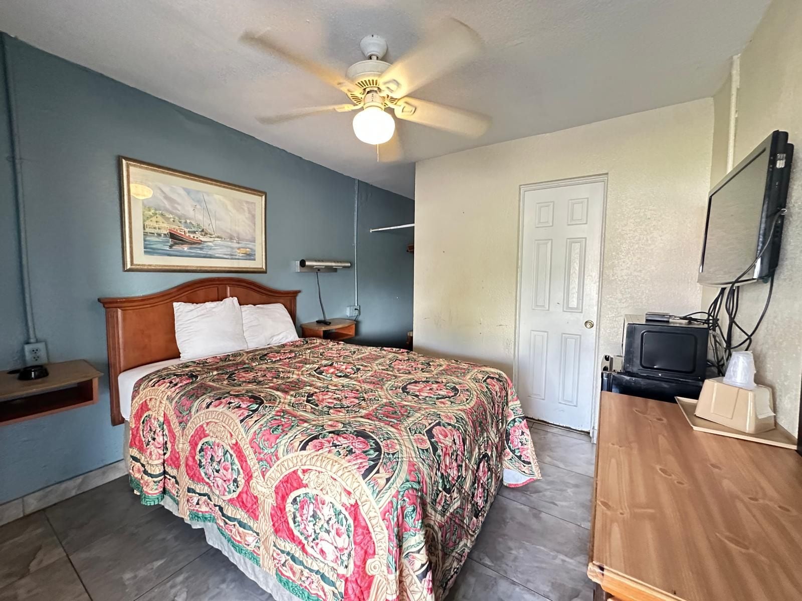 A hotel room with a bed, a ceiling fan, a painting, a television, and a door.