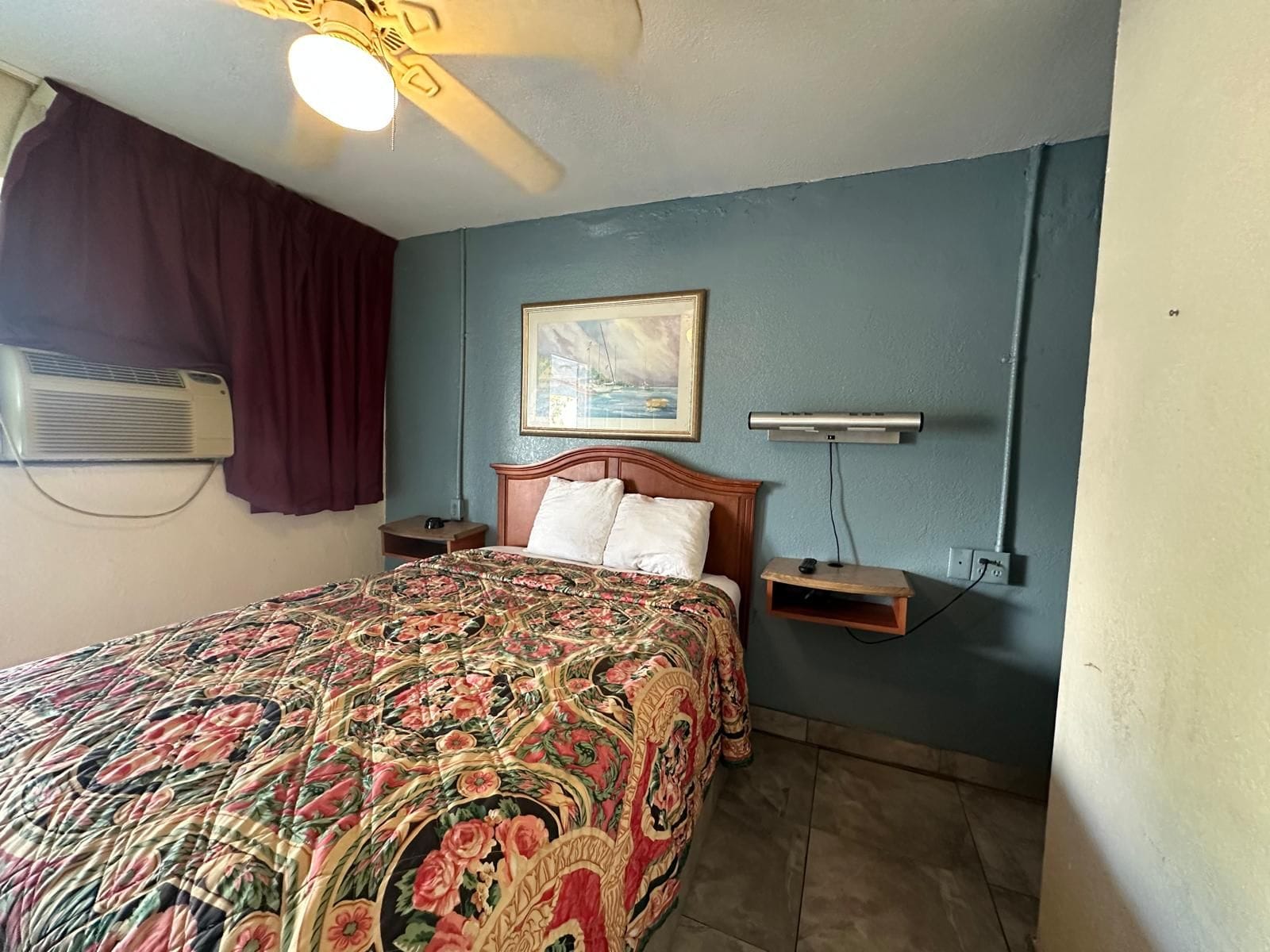 A hotel room with a bed, a ceiling fan, an air conditioning unit, a painting, and a small shelf.