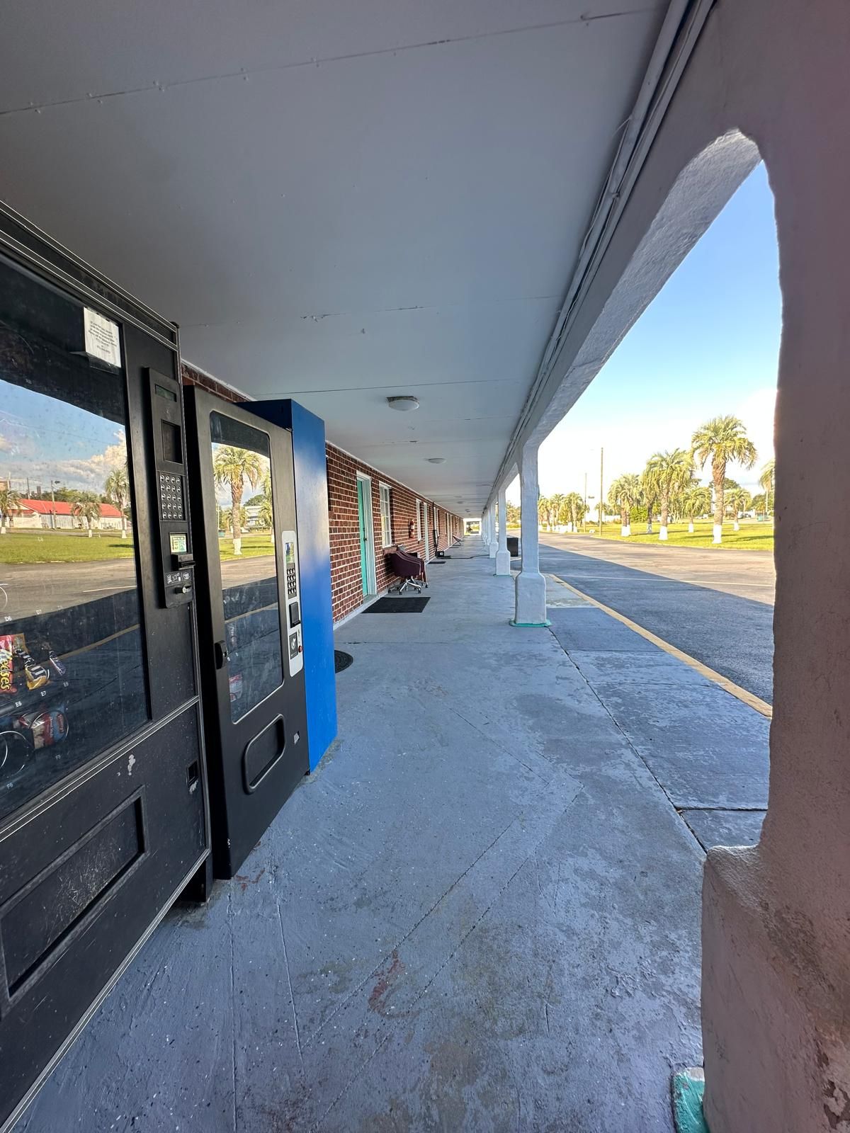 A covered outdoor area with vending machines, a bench, and a view of palm trees and a parking lot.