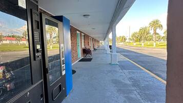 A covered outdoor area with vending machines, a bench, and a view of palm trees and a parking lot.
