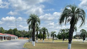 A parking lot with palm trees, a building with a red roof, and a billboard in the background. - Motel Jesup (Jesup)