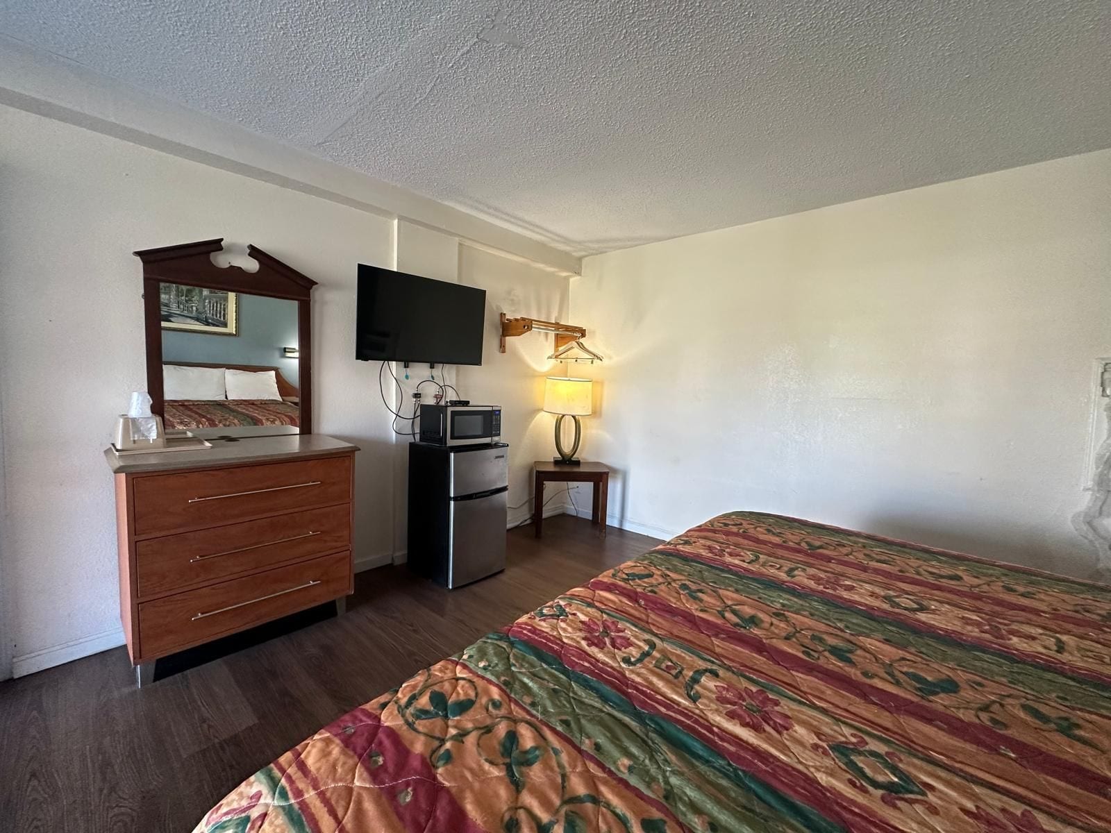 Une chambre d’hôtel comprenant un lit, une commode, un téléviseur fixé au mur, un four à micro-ondes et une petite table avec une lampe.