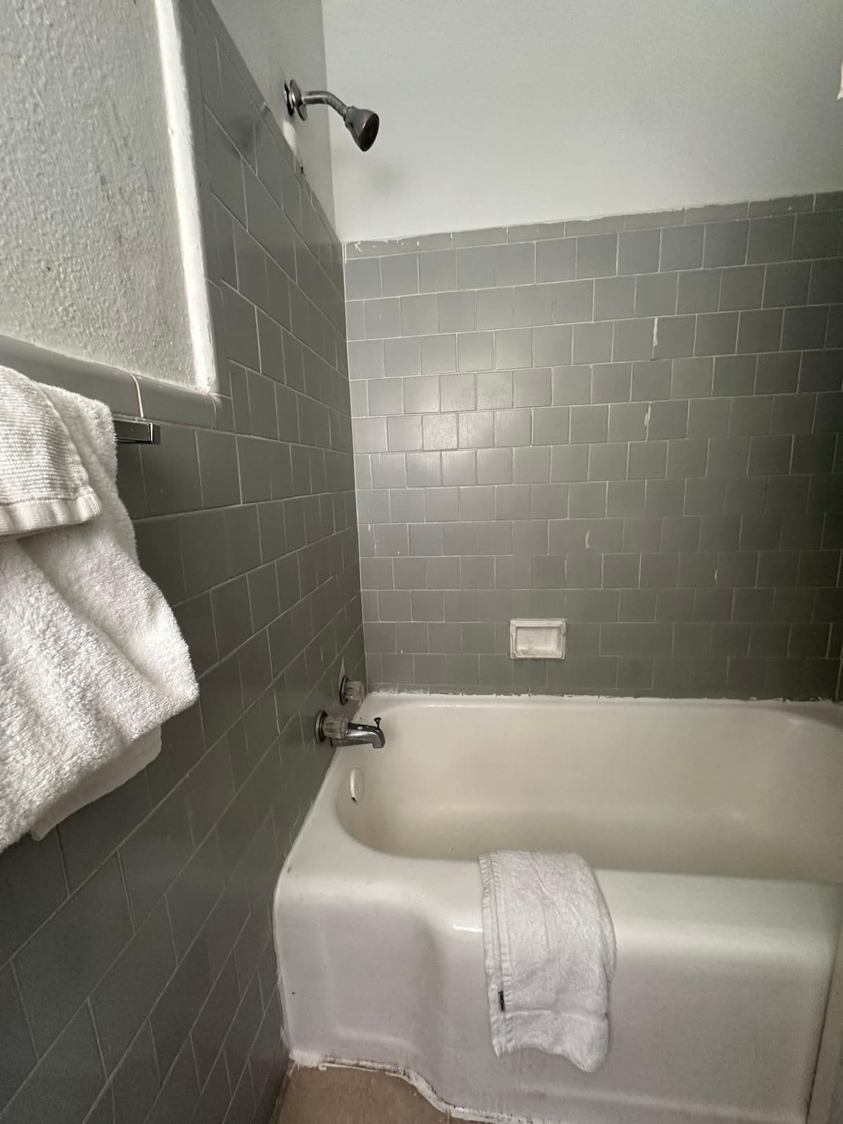 A bathroom with a bathtub, grey tiled walls, a showerhead, and a towel rack with towels.