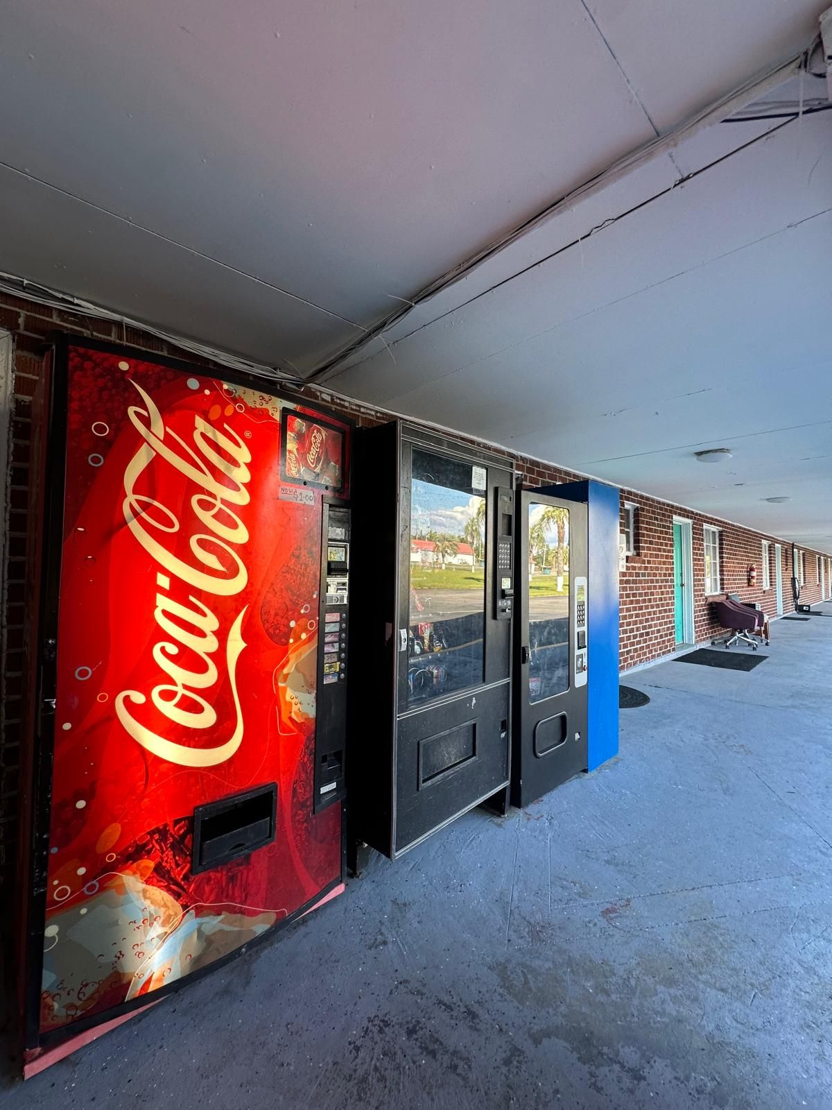 A row of vending machines with a Coca-Cola logo, a brick building, and a covered outdoor area.