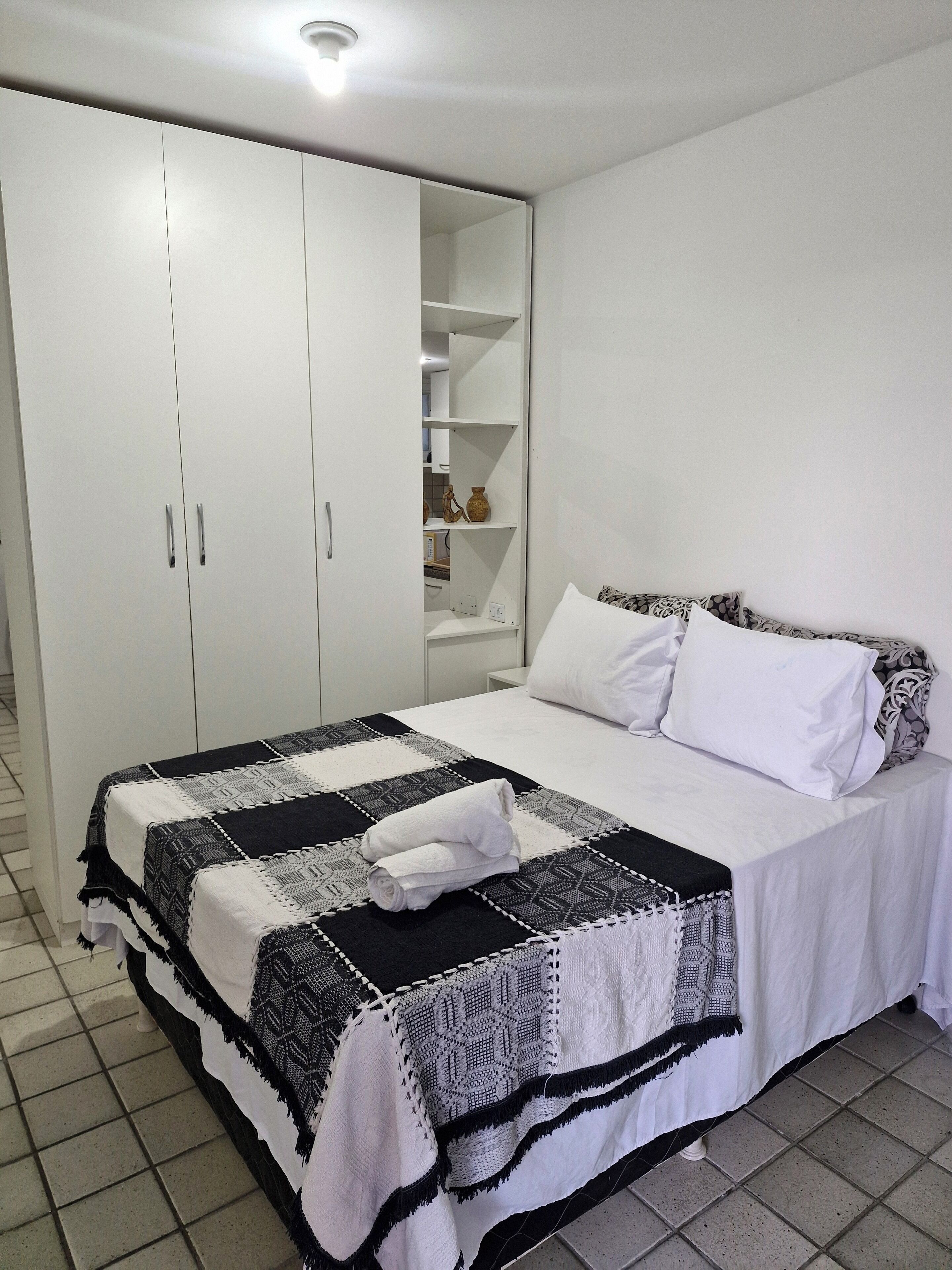 A bedroom with a bed, white curtains, a wardrobe, and a shelf with decorative items.
